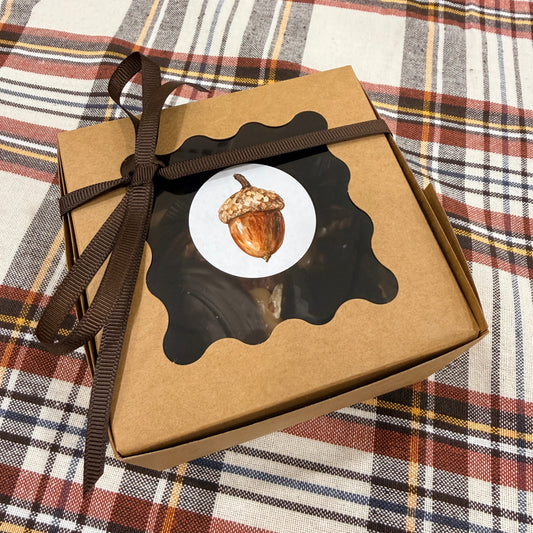 A square kraft paper gift box tied with a dark brown ribbon, featuring a decorative black scalloped label with a white circle and an acorn illustration, placed on a fall plaid blanket.